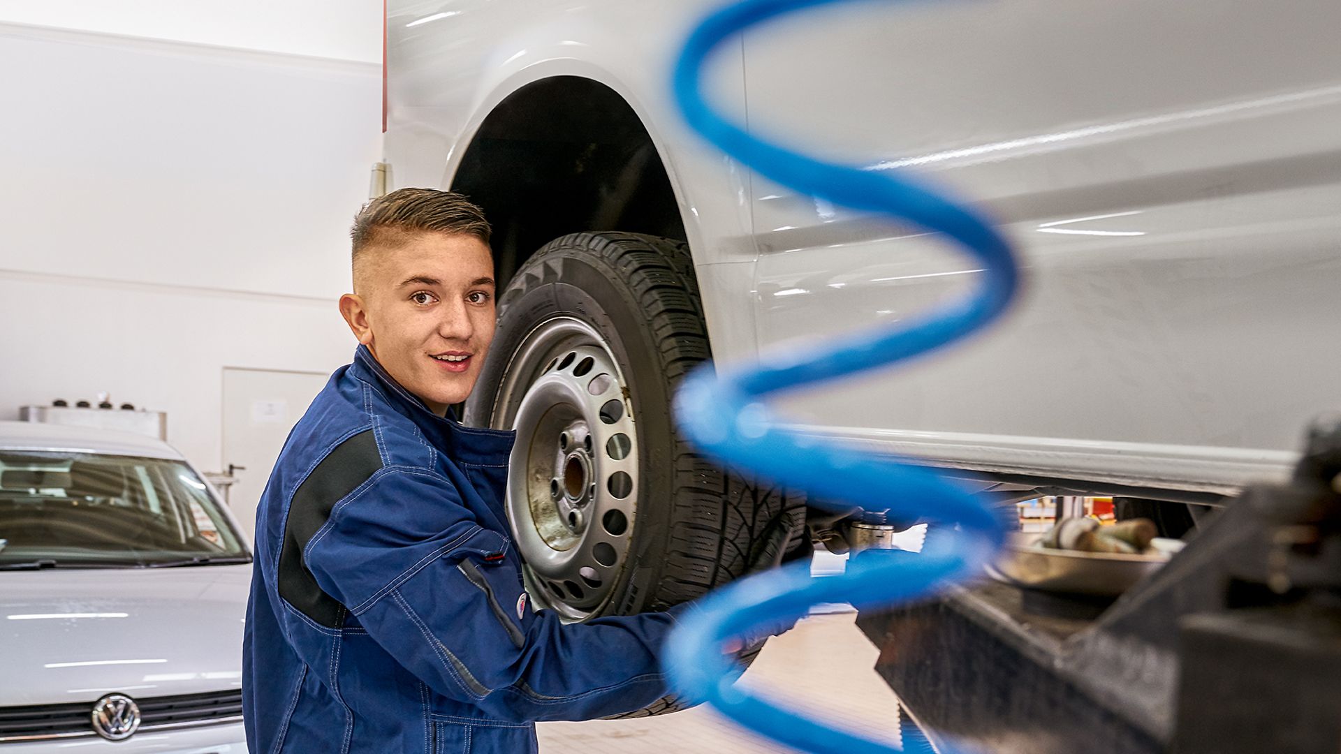 Ein Auszubildender zum KFZ-Mechatroniker steht in Arbeitskleidung neben einem aufgebockten Transporter und wechselt einen Reifen